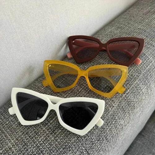 سه عینک آفتابی زنانه با فریم گربه‌ای (Cat Eye) با رنگ‌های مختلف: یک مدل سفید با شیشه تیره در جلو، یک مدل زرد کهربایی در وسط، و یک مدل قهوه‌ای تیره در بالا، که همگی روی پارچه کاناپه خاکستری قرار دارند – مجموعه عینک فانتزی فانتزی باز.