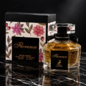 ادکلن Florence Maison Alhambra با بطری شیشه‌ای شفاف و مایع طلایی، در کنار جعبه گل‌دار مشکی.